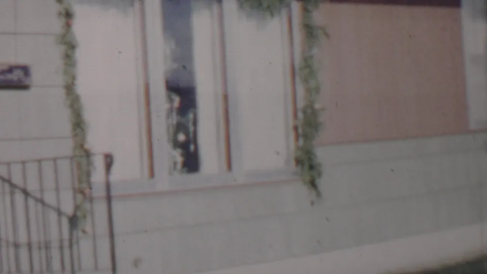 Close 1962 Lincoln, Nebraska frame of a house entry area with metal porch railings, a light-colored door, a narrow window, green garland, and a partially visible sign.