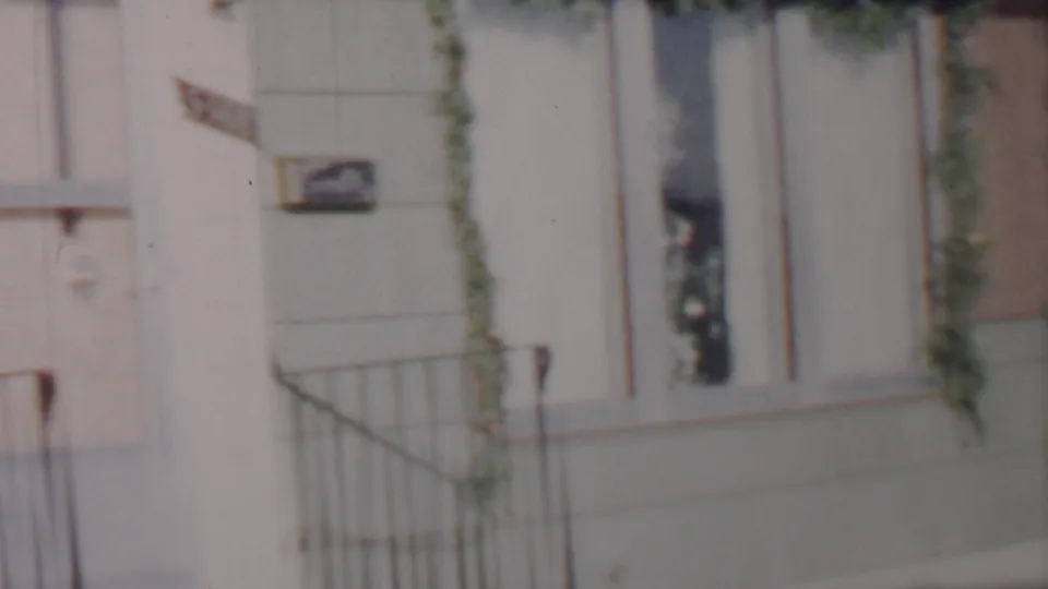 1962 Lincoln, Nebraska home-movie frame centered on the house entrance, showing the light door, metal railings, pale siding, garland near the window, and a partial sign.