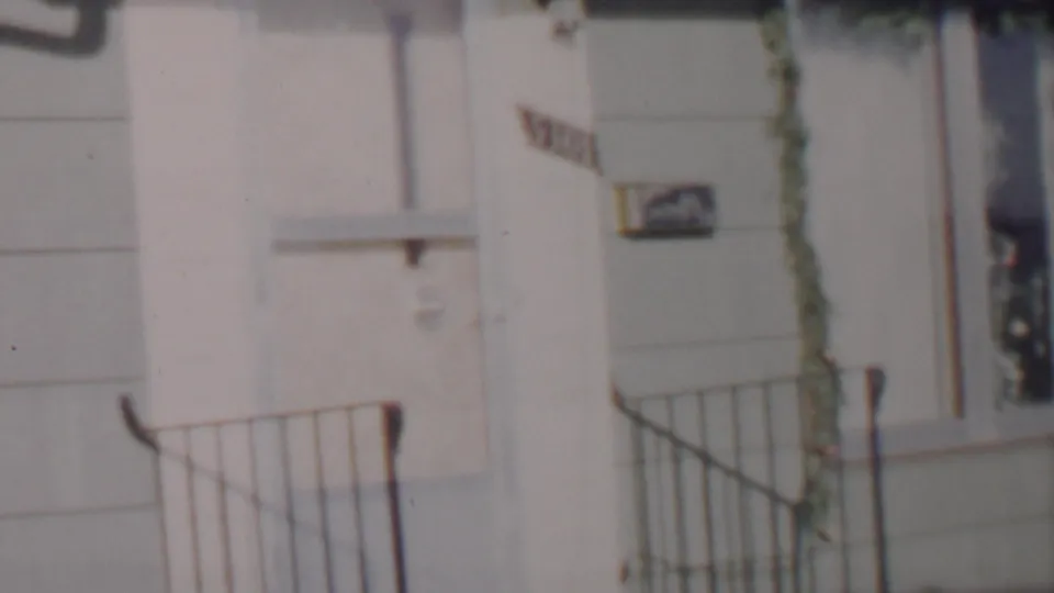 Final 1962 Lincoln, Nebraska frame showing the house corner with metal porch railing, pale siding, a narrow window edged by garland, and a partially readable sign.