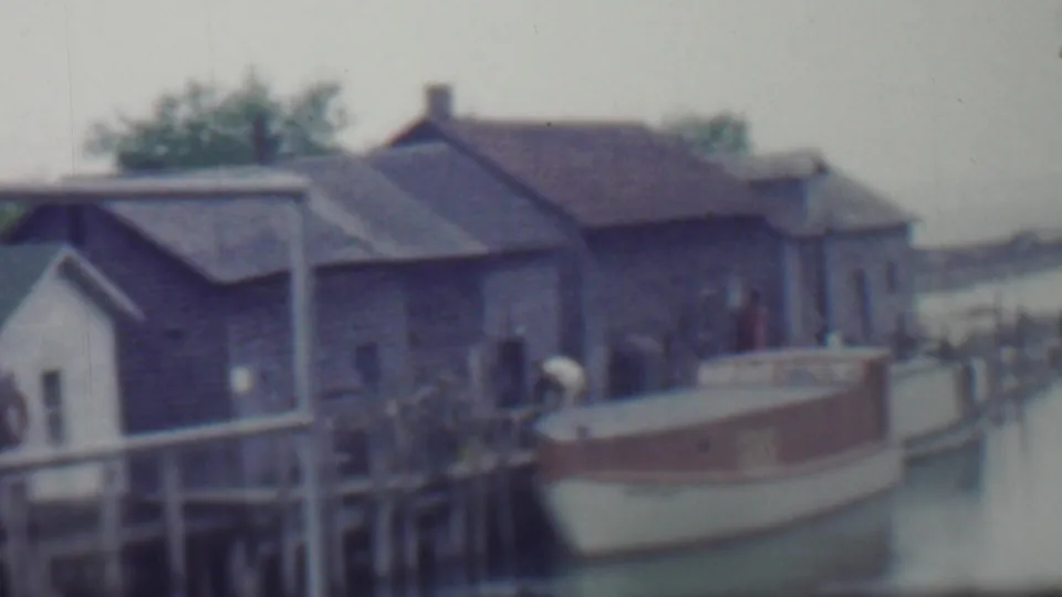 1959 Louisiana home-movie frame showing a small white and brown boat docked beside weathered wooden pilings and shingled boathouse-like structures.