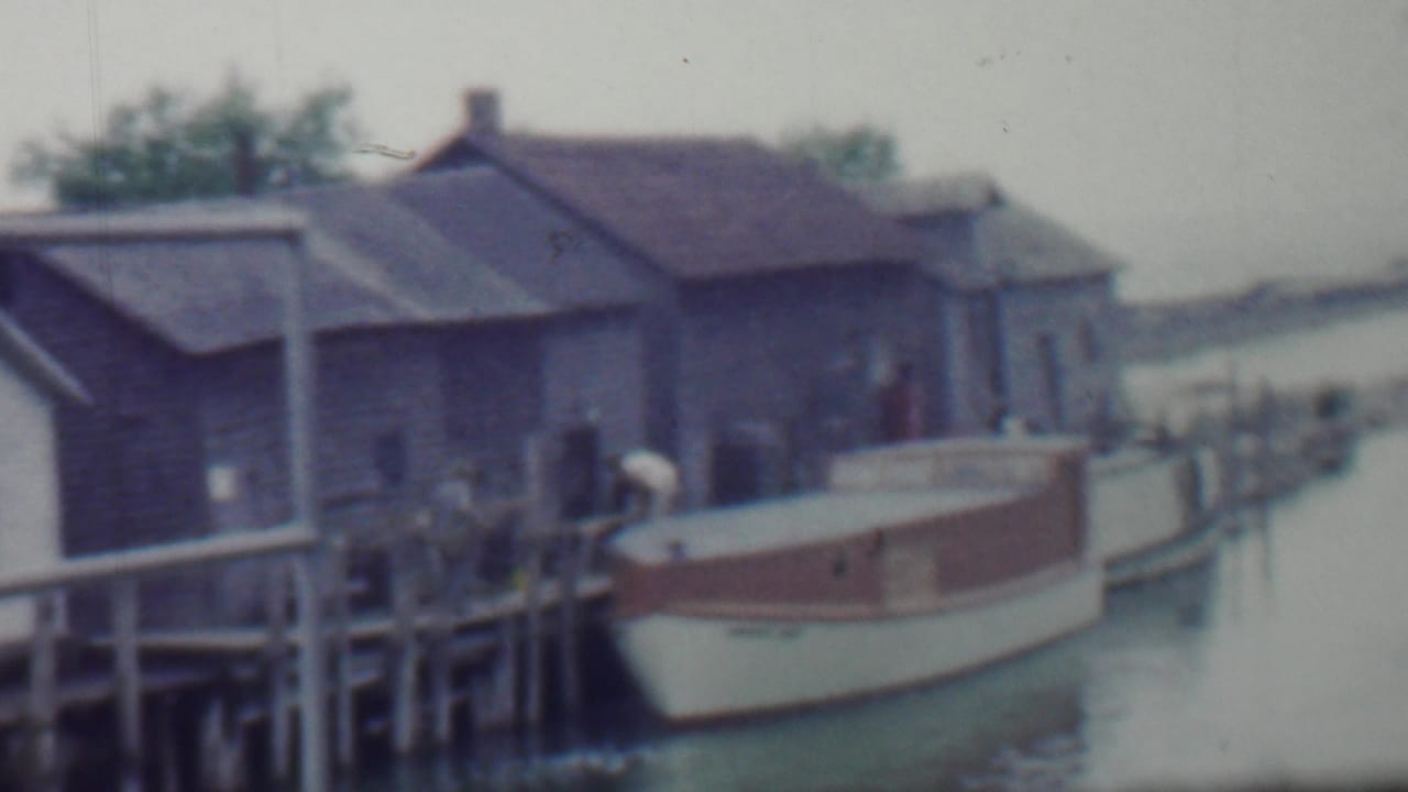 Boat moored at a wooden dock near dark shingled boathouse-like buildings.