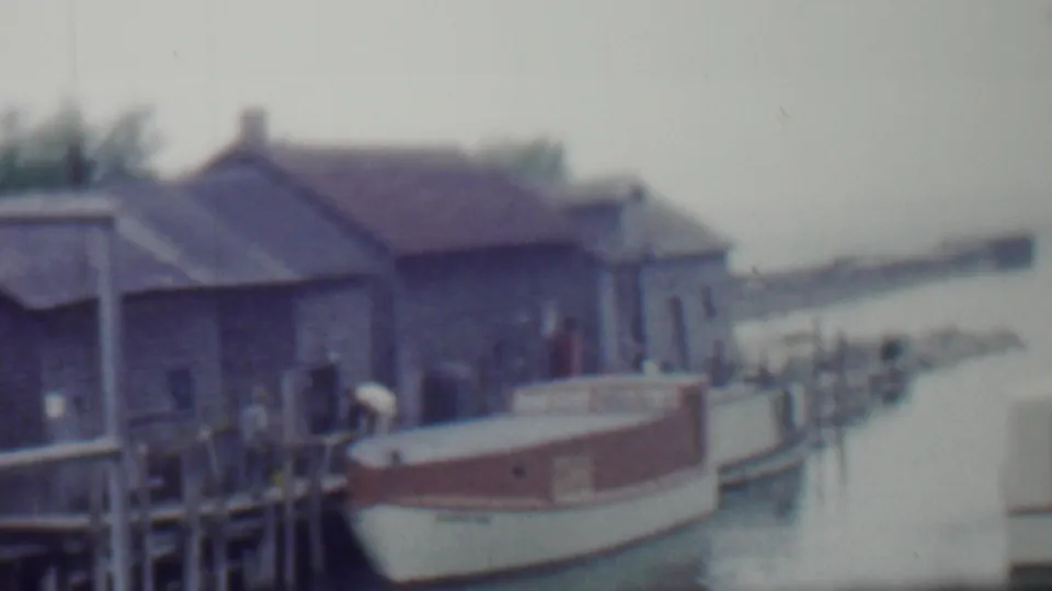 Soft 8mm frame from 1959 Louisiana showing a small painted boat alongside a wooden pier, with boathouse-like buildings and water extending behind it.