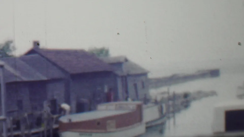 1959 Louisiana home-movie frame showing the boat, pier, wooden pilings, shingled waterfront structures, and a person standing or bending near the dock.