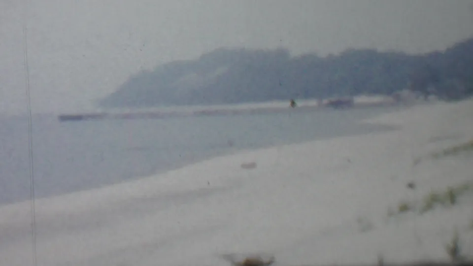 Louisiana coastal home-movie frame from supplied 1959 metadata showing a pale sandy beach, calm inlet water, and a distant tree-lined shore in soft focus.