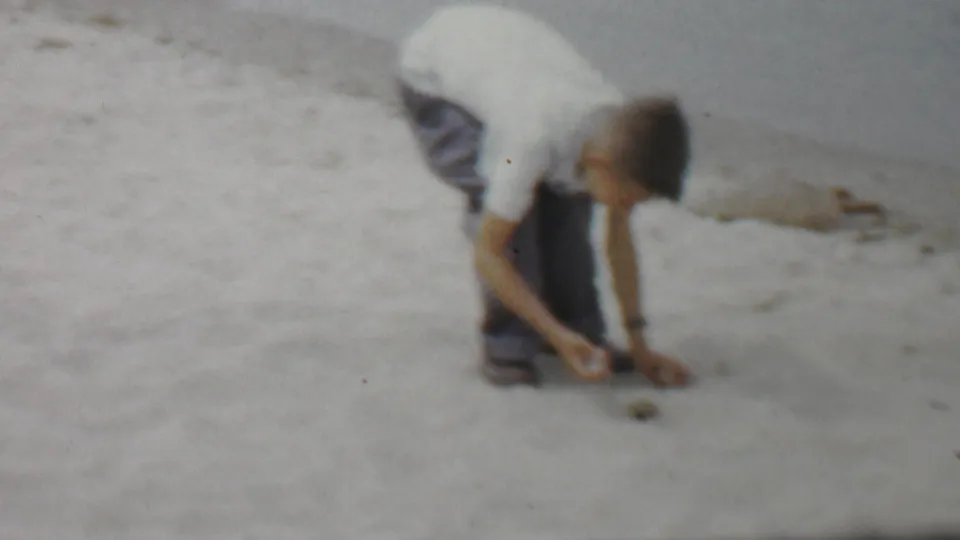 A boy in a white shirt and dark pants bends toward the sand on a Louisiana beach, with calm inlet water and a distant shoreline behind him, based on supplied 1959 metadata.