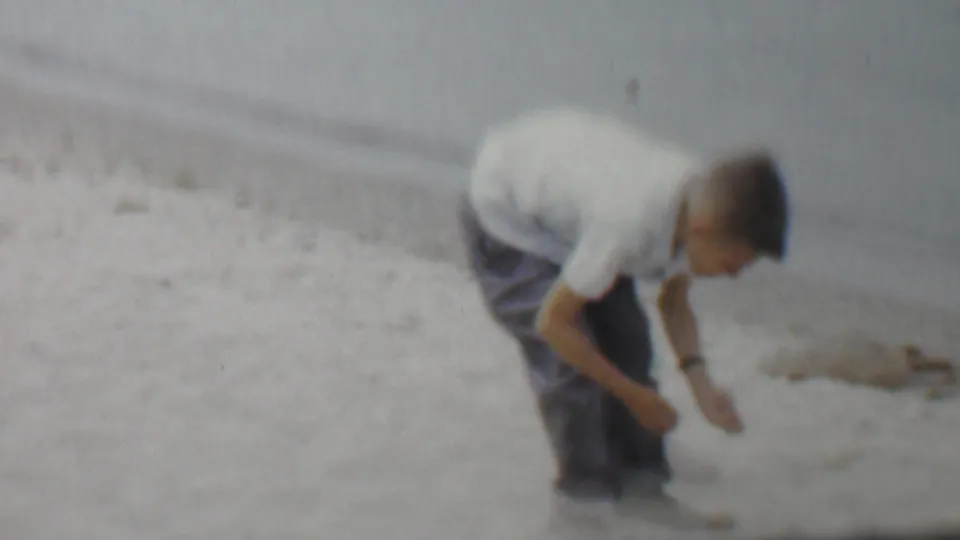 A young boy bends forward on a sandy Louisiana shoreline near calm water, with the soft focus and faded color of supplied 1959 8mm home-movie footage.