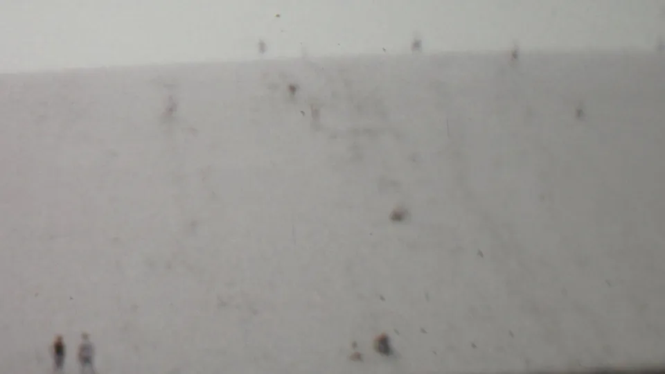A faded 8mm frame from the supplied 1959 United States clip shows distant people dispersed across a soft, low-contrast hillside.