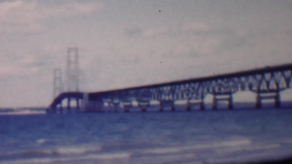Soft 8mm frame from the supplied 1959 Louisiana clip showing distant bridge towers, a truss span, and rippling water under an overcast sky.