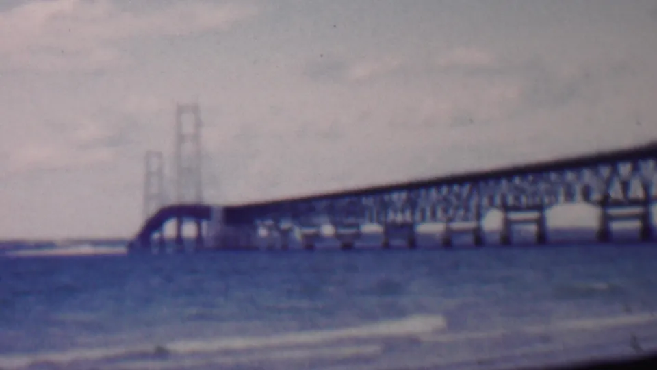 1959 Louisiana home-movie still showing a bridge crossing choppy water, with pale clouds, softened focus, and faded blue-purple film color.