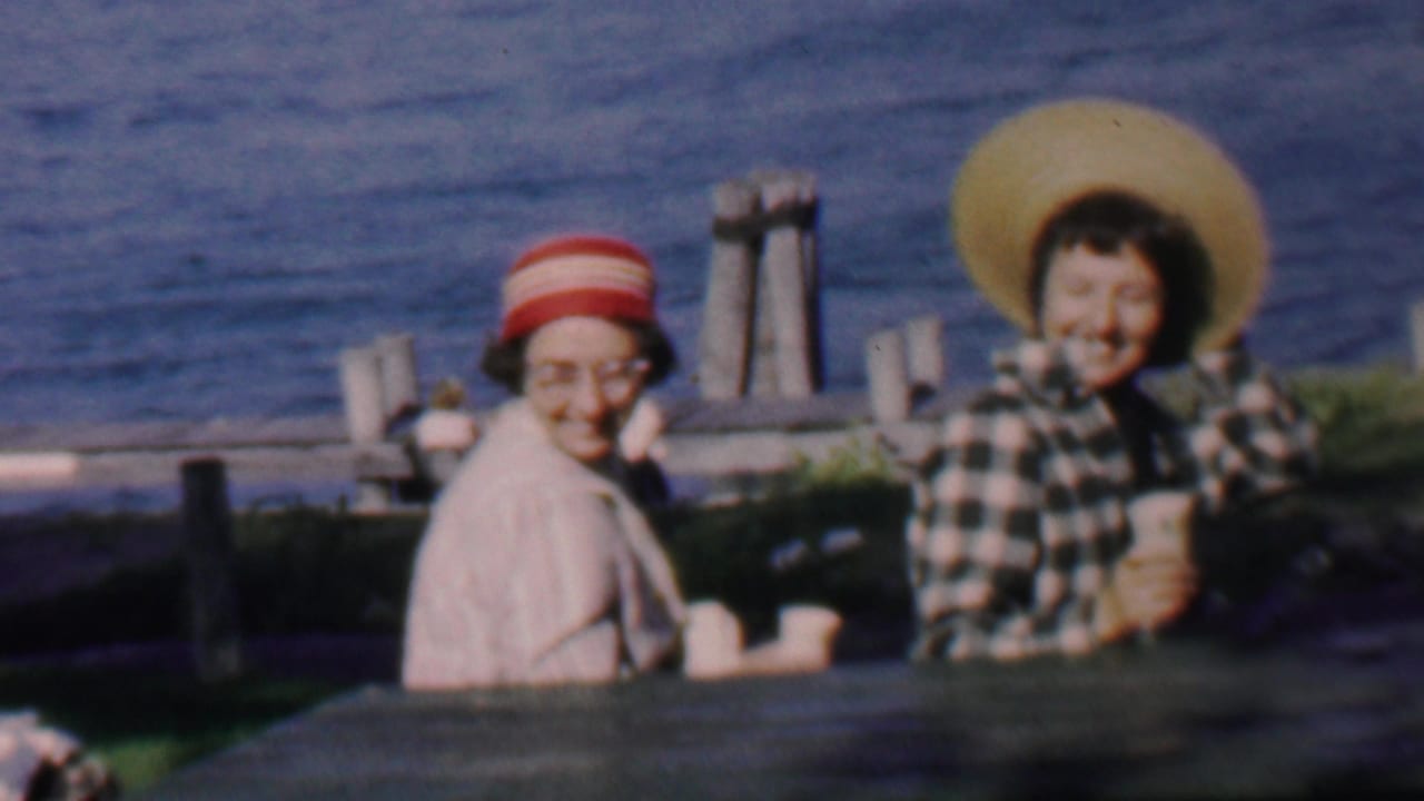 In 1959 Louisiana home-movie footage, two adult women sit at a wooden picnic table by open water, one in a striped hat and glasses and the other in a wide straw hat.