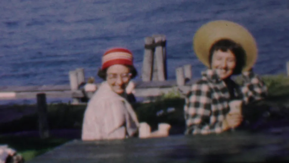 In 1959 Louisiana home-movie footage, two adult women sit at a wooden picnic table by open water, one in a striped hat and glasses and the other in a wide straw hat.