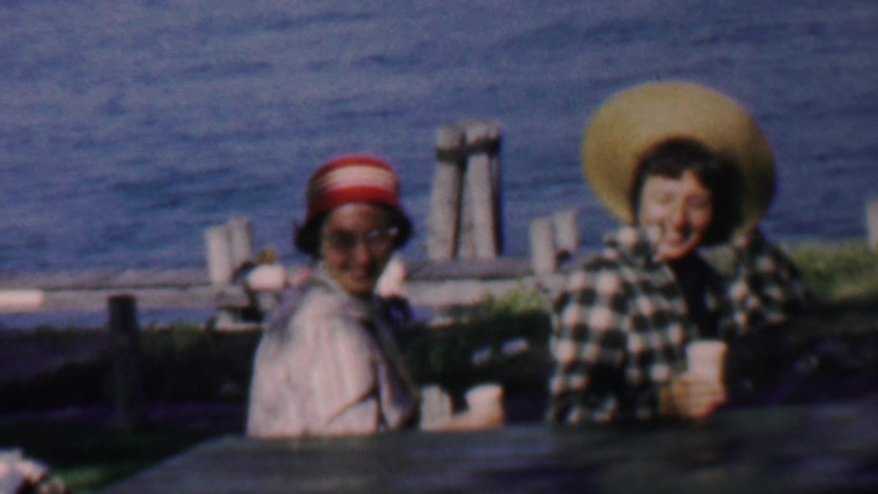 The two women remain seated by the water in the 1959 Louisiana home-movie scene, with paper cups visible on the table and dock-like posts in the background.