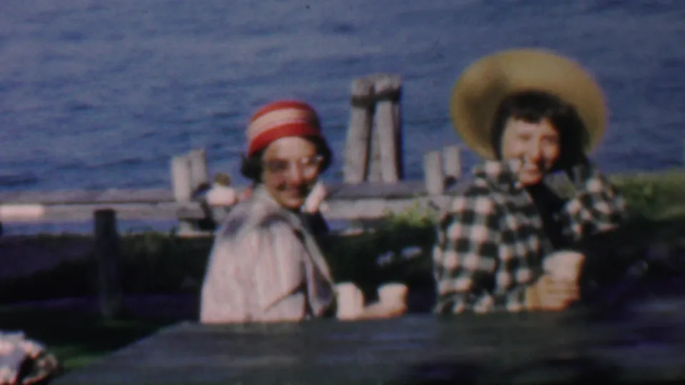 A soft 8mm frame shows two adult women smiling at the camera from a waterfront picnic table, with the straw hat, striped hat, cups, and water still clearly visible.