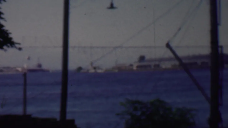 1959 Louisiana home-movie frame showing a distant waterfront across dark blue water, with boats, a low shoreline structure, utility poles, wires, and foliage in the foreground.