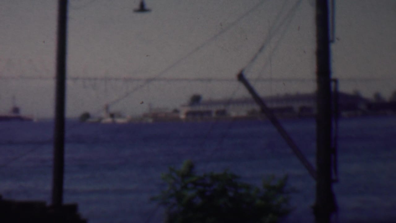 1959 Louisiana 8mm frame looking across water toward boats and a low building or pier, with dark utility poles and diagonal wires interrupting the foreground.