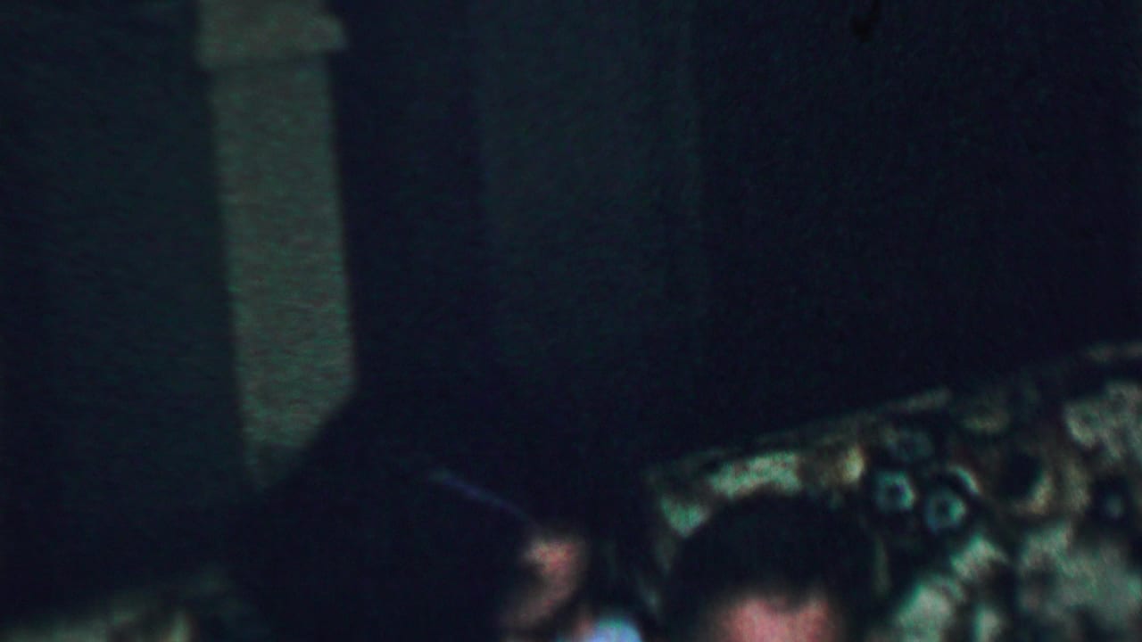 A low-light 1959 home-movie frame from the United States shows part of a seated woman near floral-patterned upholstery in a private living room.