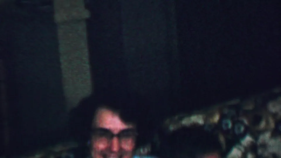 A woman with dark hair and glasses smiles while seated near floral-patterned upholstery in a dim 1959 American home-movie interior.