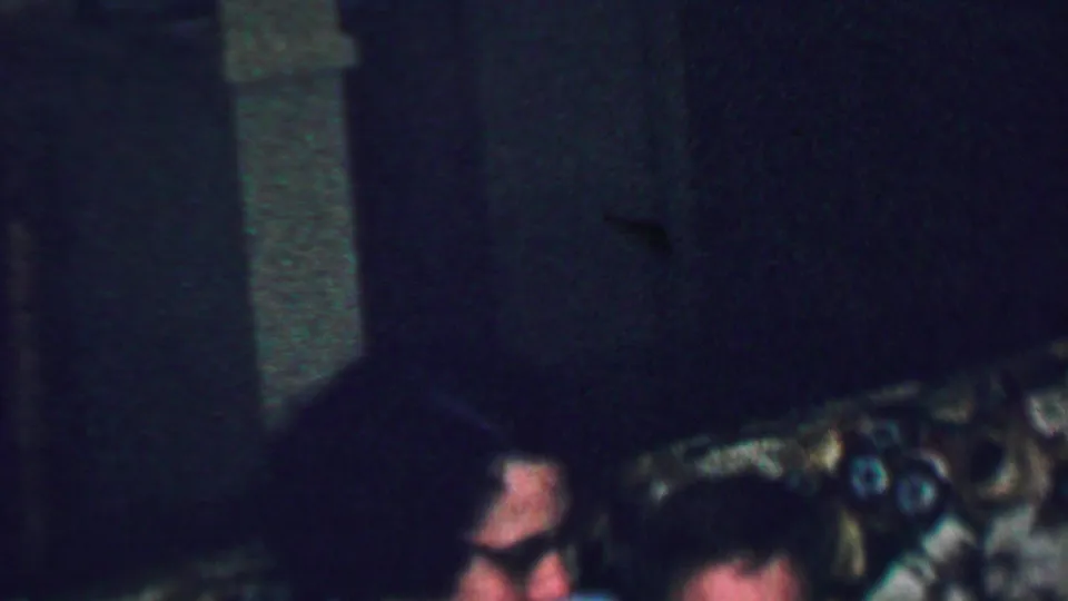 A dark, grainy 8mm frame shows partial faces of seated family members near patterned living-room upholstery, with the main woman lower in frame.