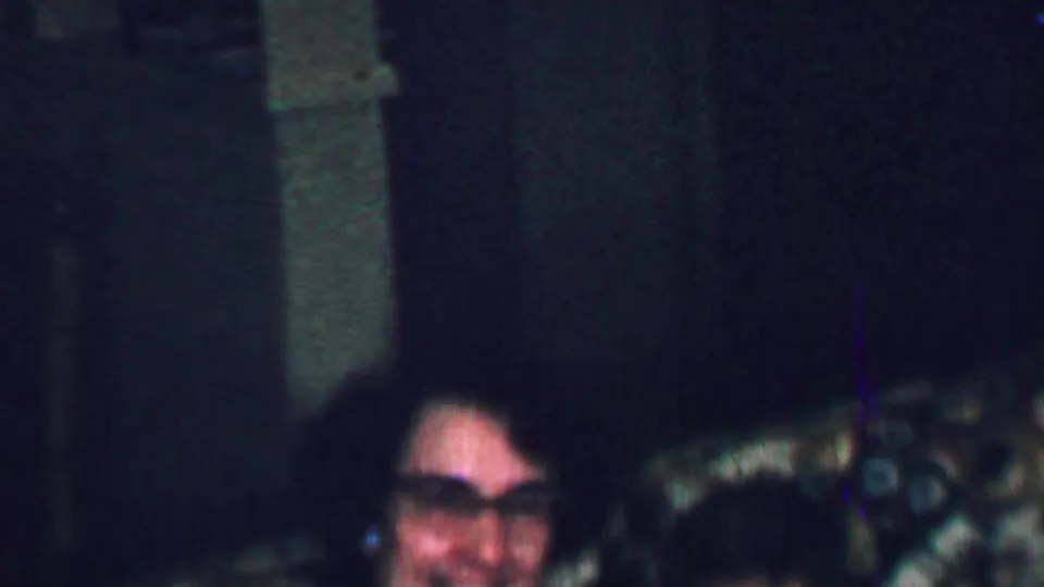 A smiling woman with glasses sits beside another partly visible person on patterned seating in a dim 1959 United States home-movie living room.