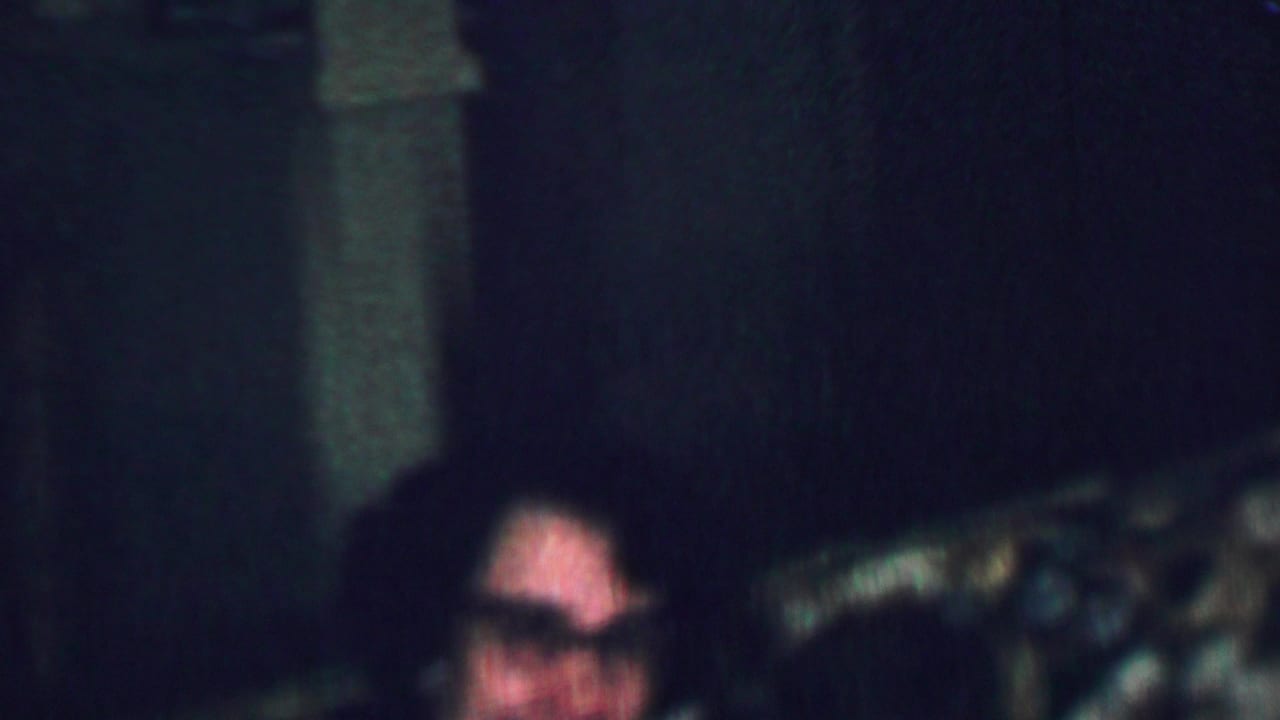 In supported 1959 Omaha home-movie footage, a dark-haired woman wearing glasses smiles while seated in a low-lit domestic interior.