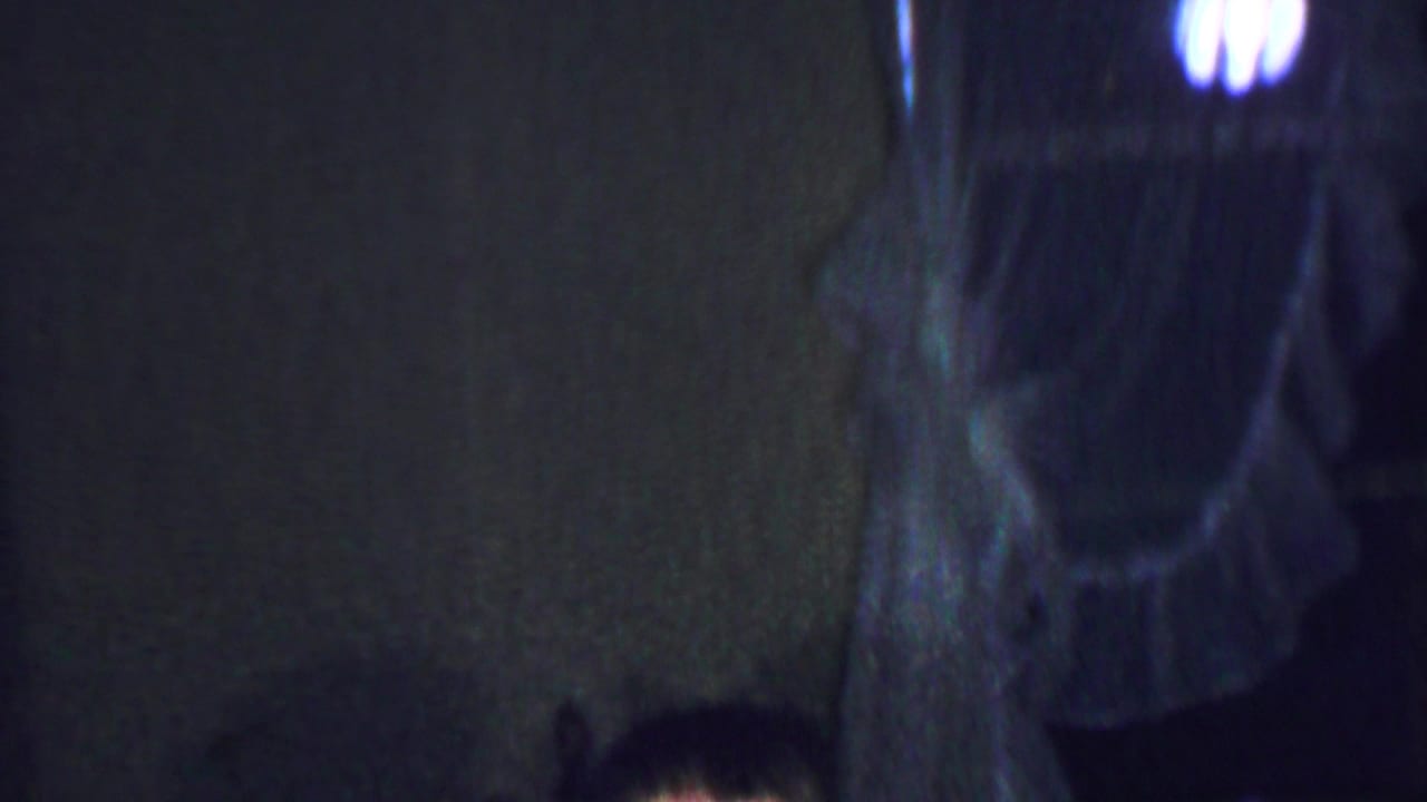 A low-light 8mm frame shows the surrounding room, including textured wall surfaces, dark curtains, and a bright out-of-focus light source.