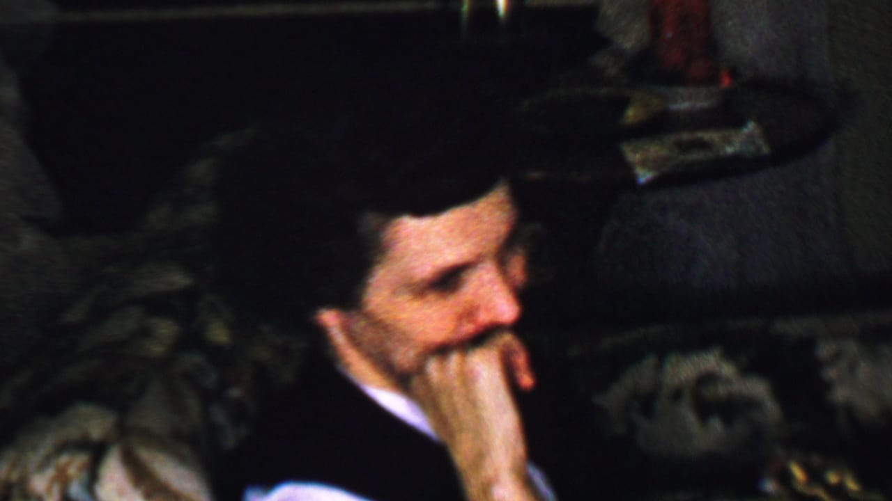 In this Omaha, Nebraska home-movie frame from the supplied 1959 metadata, a mustached adult man in a dark suit and white shirt sits on a patterned couch in dim interior light.
