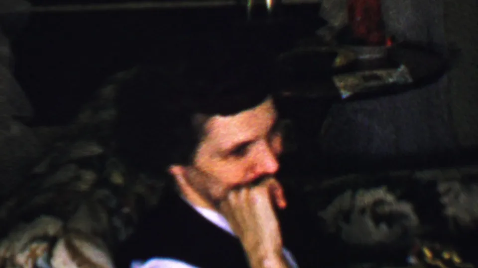 In this Omaha, Nebraska home-movie frame from the supplied 1959 metadata, a mustached adult man in a dark suit and white shirt sits on a patterned couch in dim interior light.