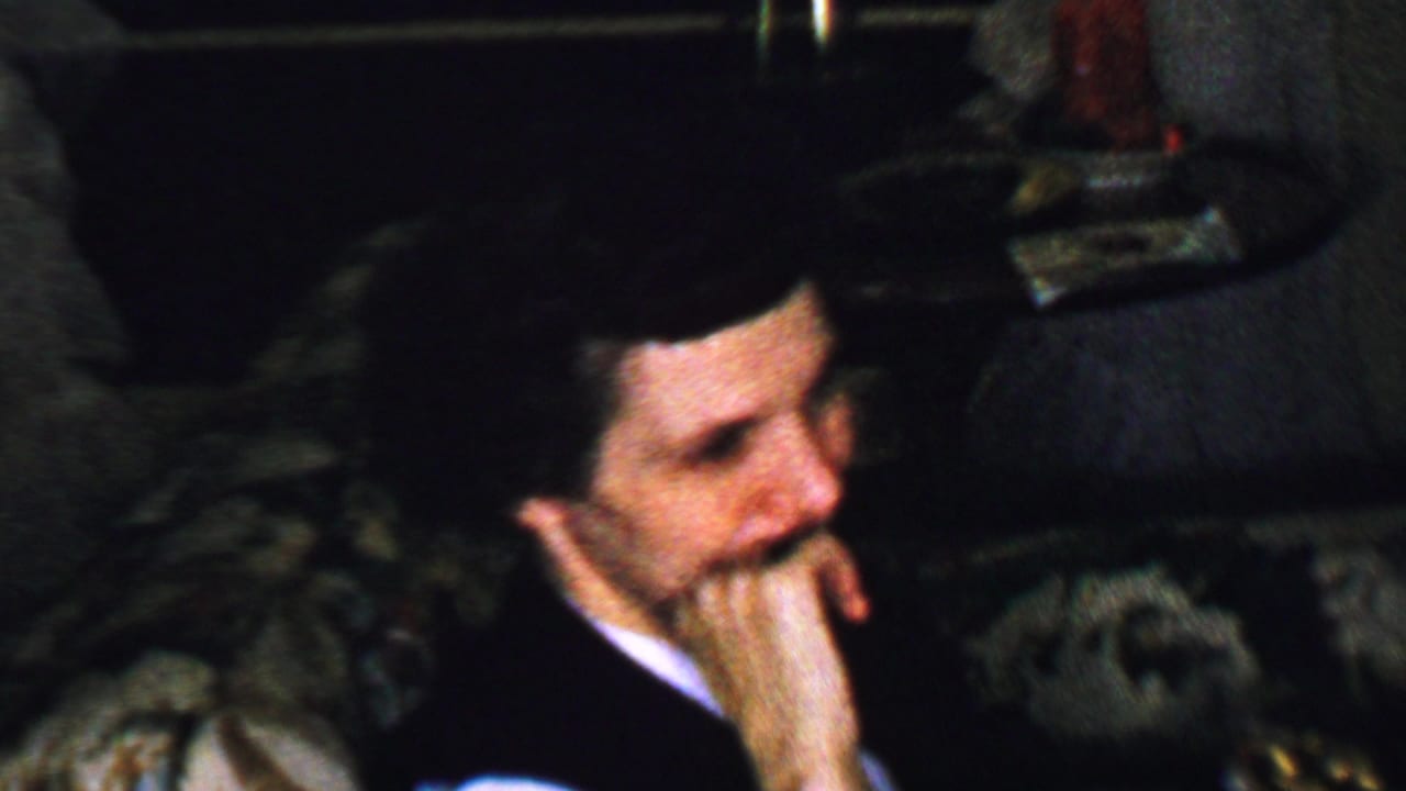 Mustached man in a dark suit sits on a couch with his hand against his face.