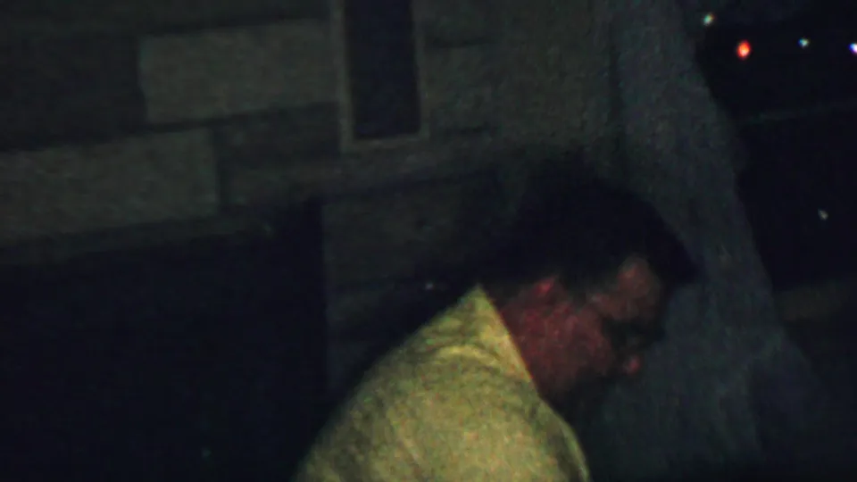 A different adult man wearing glasses and a light-colored shirt is shown in profile within the same dark private interior, partly obscured by low light and film grain.