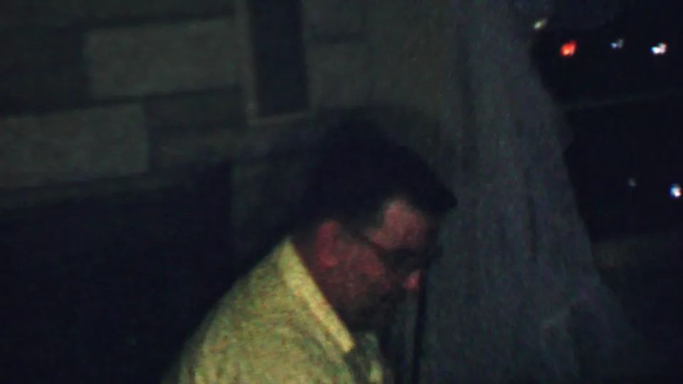 The man in glasses remains seated in profile in the dimly lit room, with his light shirt and face partly softened by the low-light 8mm exposure.