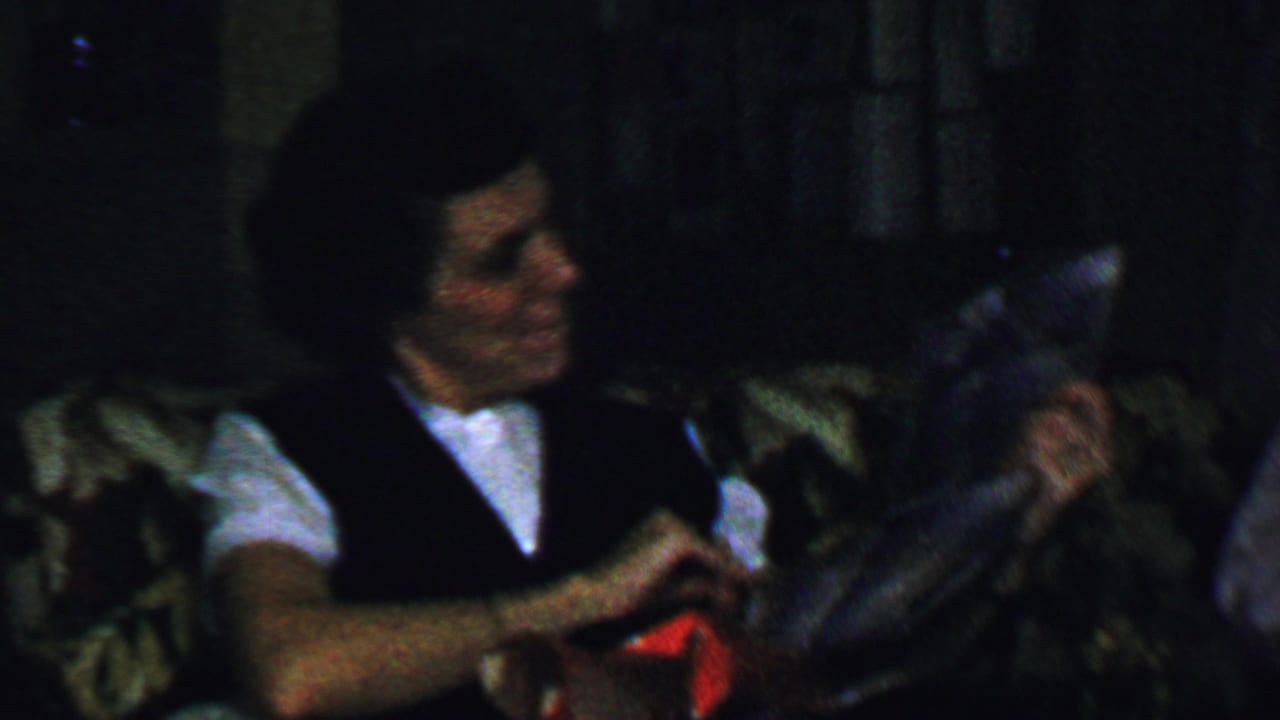 In a 1959 Omaha home-movie frame, a woman in a white shirt and dark vest sits on a patterned couch while holding or unwrapping a gift.