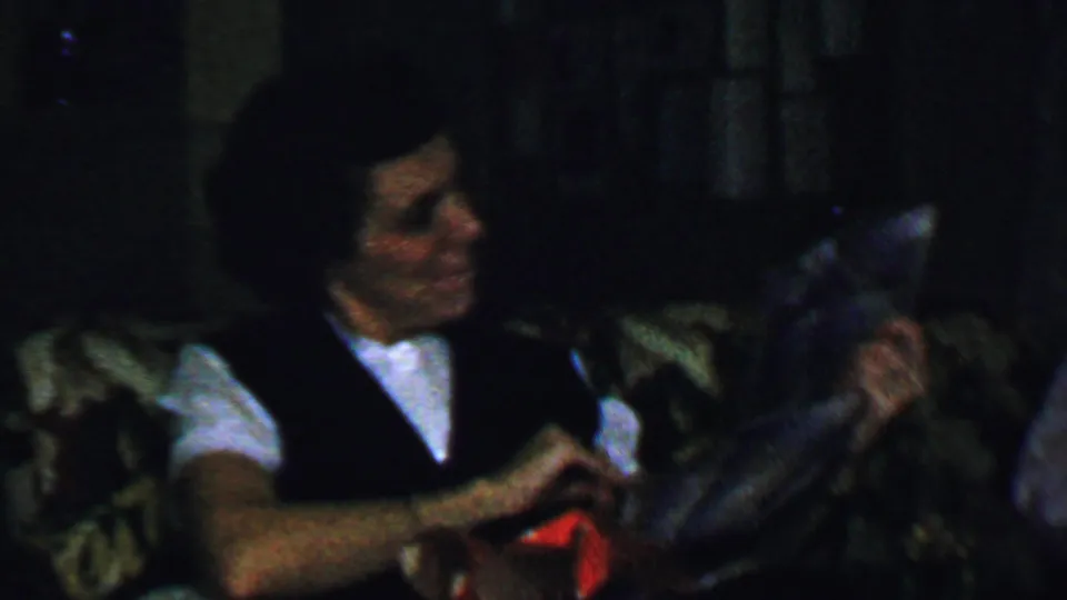 In a 1959 Omaha home-movie frame, a woman in a white shirt and dark vest sits on a patterned couch while holding or unwrapping a gift.