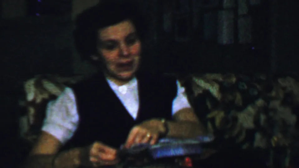 The seated woman looks down toward her hands as she continues inspecting the gift or its packaging in the living room.