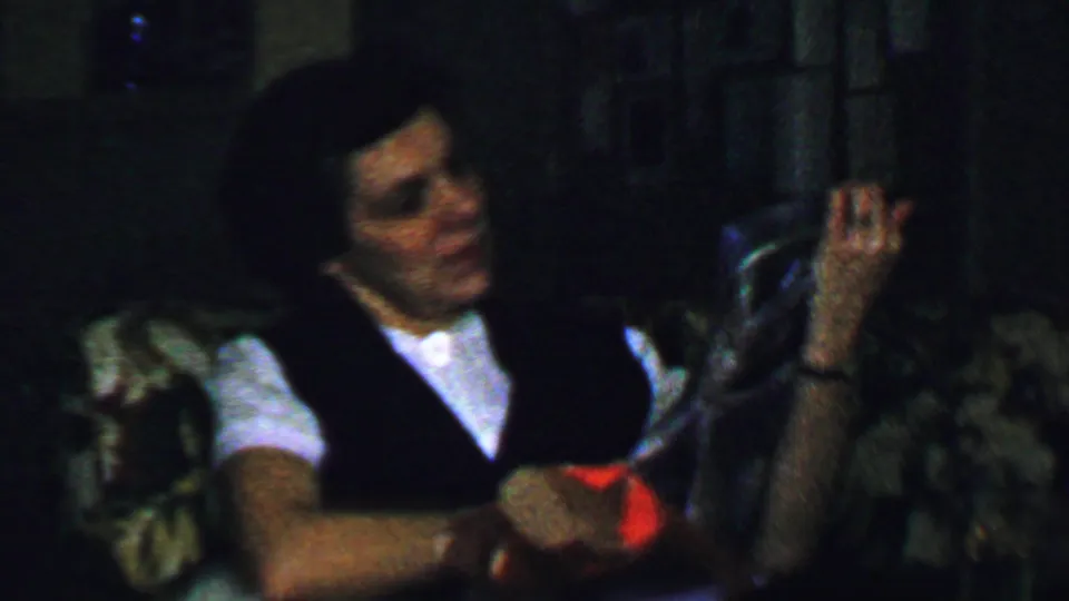 The woman raises a small object or piece of packaging from the gift while seated on the patterned couch in the Omaha home-movie scene.