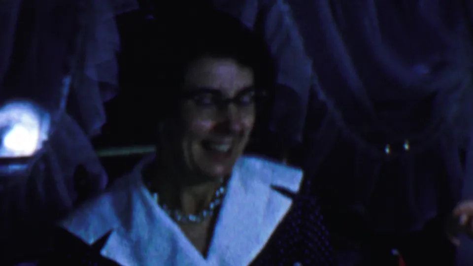 In supported 1959 Omaha home-movie footage, a woman in glasses, a polka-dot dress with a white collar, and a necklace smiles in a dim private interior.