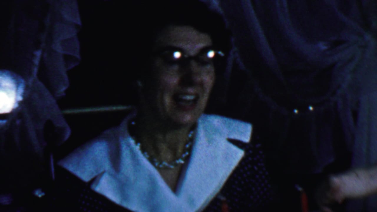 Woman in glasses smiles broadly beside standing people in low light.