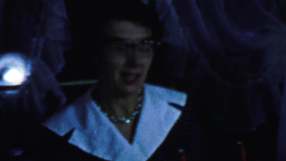 A soft-focus 8mm frame shows the seated woman looking down or toward the table, with her white collar, glasses, and surrounding figures still visible.