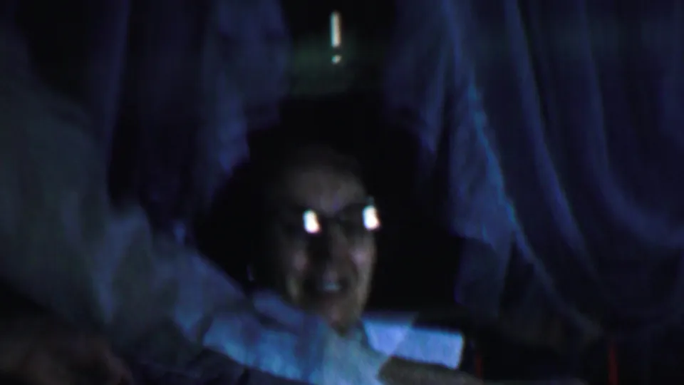 At the end of the clip, the woman smiles from the lower right of the dim 1959 home-movie frame while standing people and draped fabric fill the background.