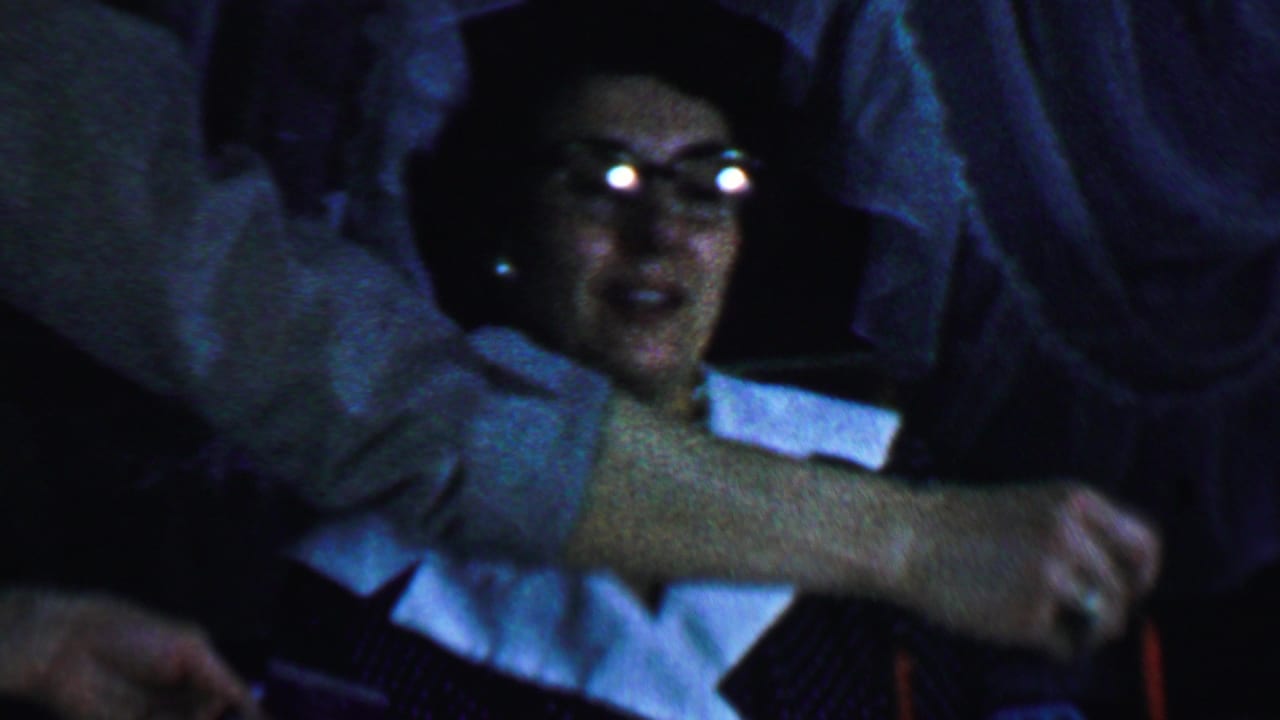 In a 1959 Omaha home-movie frame, an older woman wearing glasses, a dark dress with a white collar, and a necklace sits near a red candle while a hand reaches across the foreground.