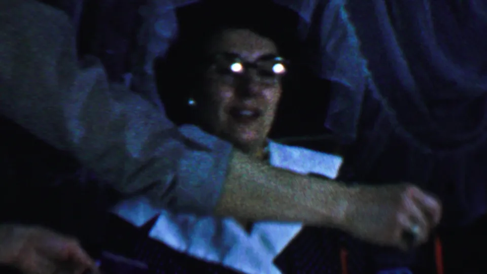 In a 1959 Omaha home-movie frame, an older woman wearing glasses, a dark dress with a white collar, and a necklace sits near a red candle while a hand reaches across the foreground.