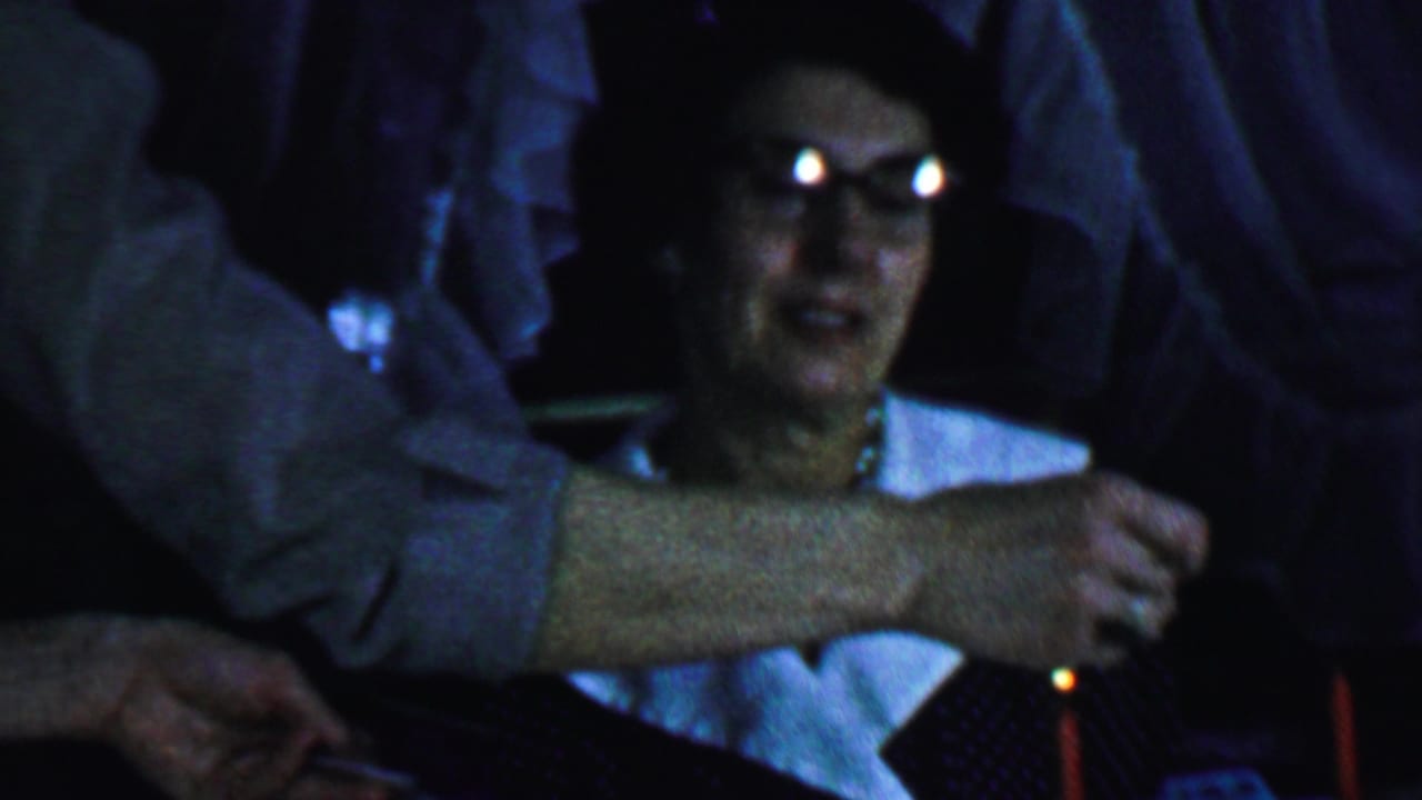 Older woman smiles in low light while an arm reaches toward a candle in front of her.