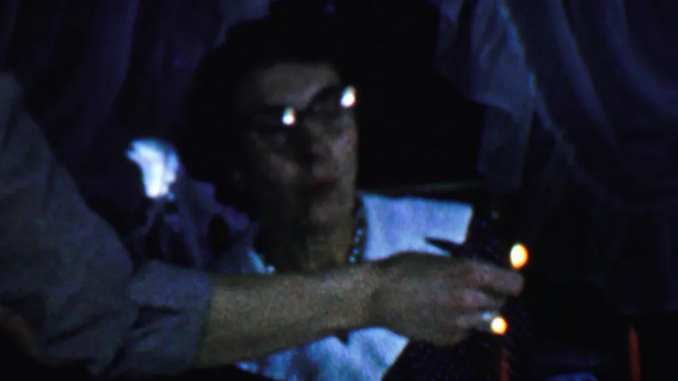The frame shows the woman partly hidden by a foreground arm while a small candle flame glows near her dark polka-dot dress and white collar in the dim birthday scene.