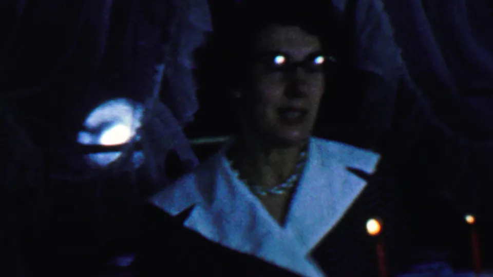 At the end of the clip, the older woman looks off to the side beside red candles, her glasses catching light above a white-collared dress and pearl necklace.