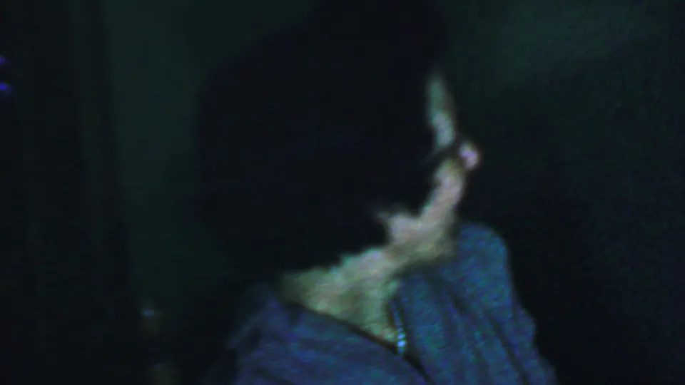 In this supported 1959 Omaha home-movie frame, a woman with glasses and dark curly hair appears in profile against a dim, grainy background.