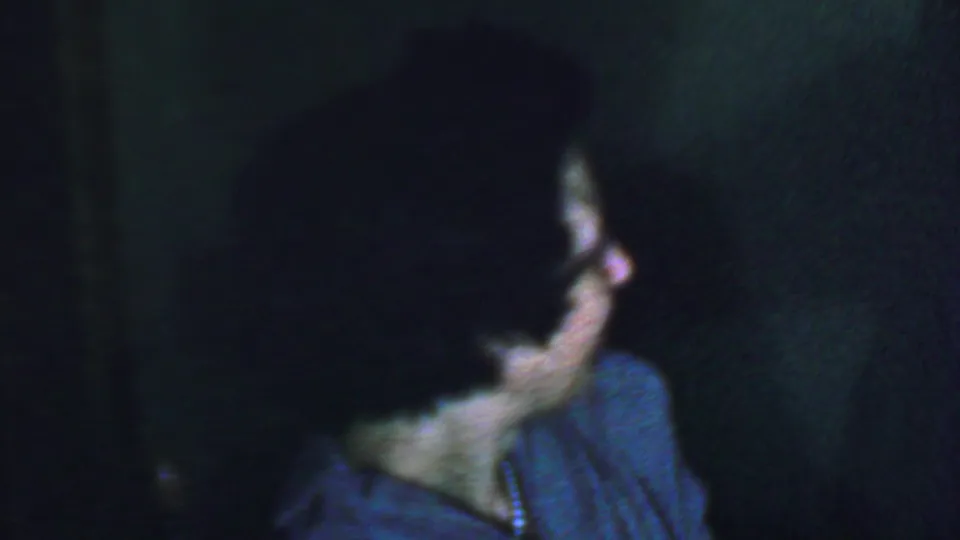 The frame shows a soft-focus profile of a woman with glasses, dark curly hair, and a blue-gray garment in low-light 8mm footage.