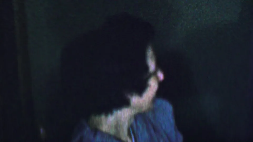 A woman in glasses remains in profile near the end of the clip, with her blue-gray top and dark hair visible in grainy low-light 8mm footage.