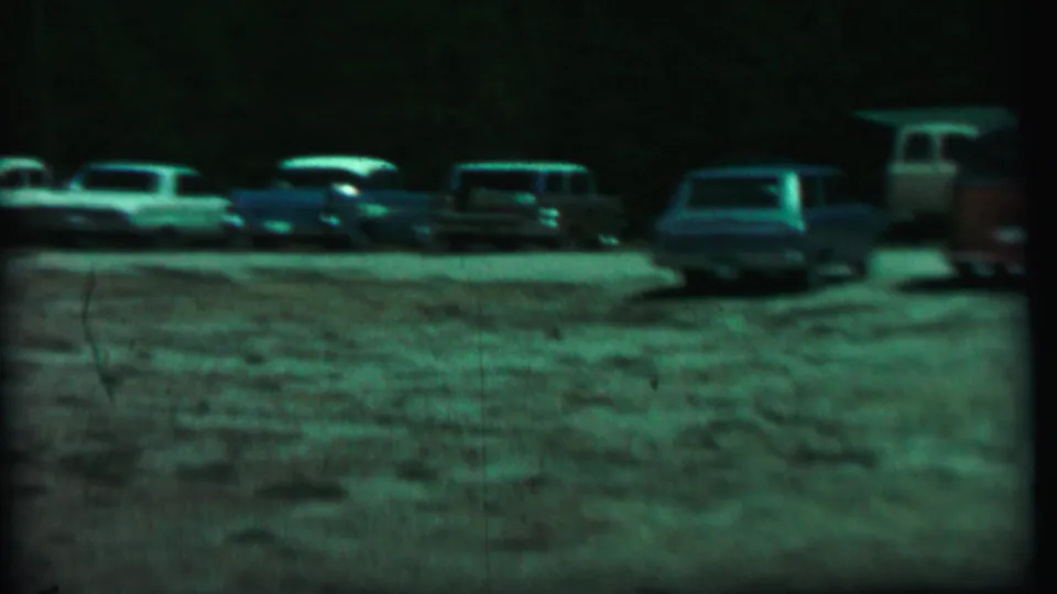 1967 home-movie frame from Farragut State Park showing several parked cars on a gravel lot under dark, green-tinted exposure.