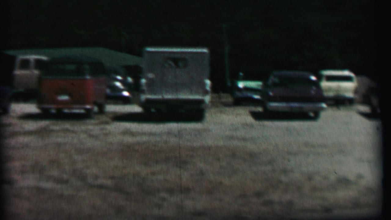 1967 home-movie frame from Farragut State Park showing a gravel parking lot with a red station wagon, light utility truck, sedans, and dark trees in the background.