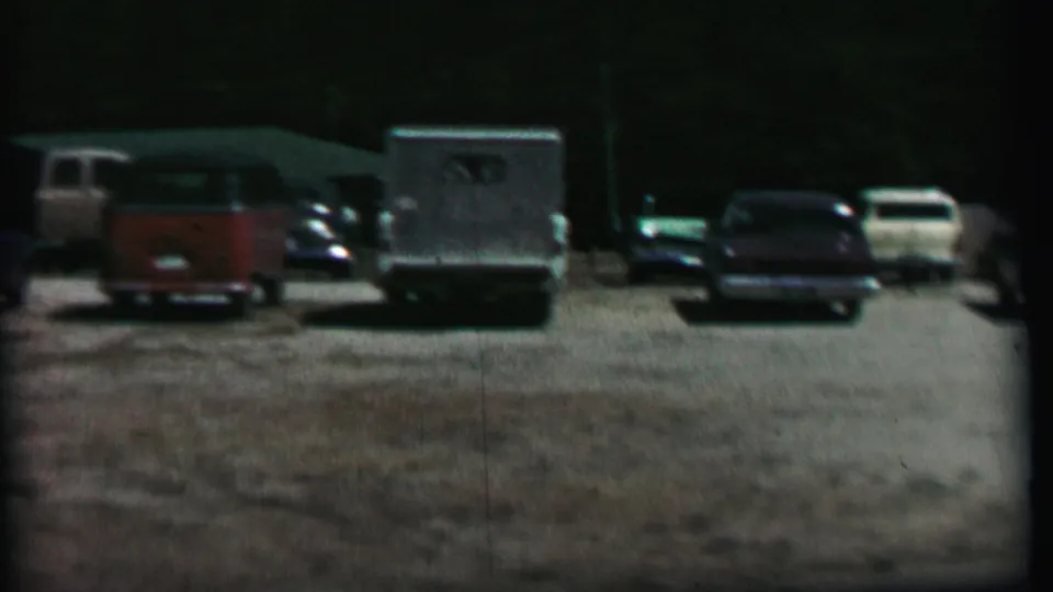 1967 home-movie frame from Farragut State Park showing a gravel parking lot with a red station wagon, light utility truck, sedans, and dark trees in the background.