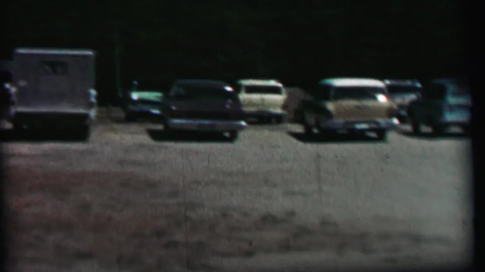 1967 home-movie frame at Farragut State Park showing several parked automobiles and a boxy utility vehicle across a dim gravel parking area.